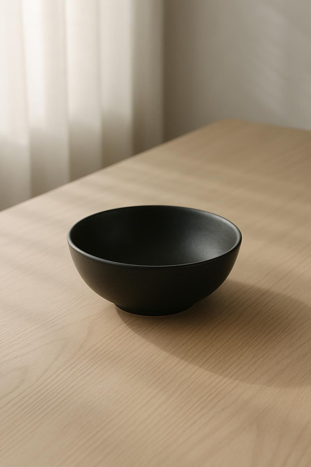 A single matte black ceramic bowl with gently curved edges sits centered on a smooth, pale oak tabletop. The entire scene is illuminated by soft, filtered daylight streaming through an unseen sheer curtain, casting subtle, elongated shadows and delicate gradients along the surface. The minimalist composition is free from clutter, focusing solely on the refined simplicity of the bowl and the organic grain of the tabletop. The overall mood is calm and contemplative, with an understated sophistication that underscores the elegance of minimalism. Captured from a slightly elevated angle with sharp focus on the bowl and a gentle falloff into the softened background, evoking a sense of peaceful order and clarity. The photographic style is clean, muted, and meticulously composed to reflect a sophisticated minimalist aesthetic.